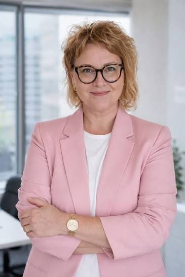 Kobieta w różowej marynarce i okularach, stoi z założonymi rękami w jasnym biurze. Jest uśmiechnięta i pewna siebie.