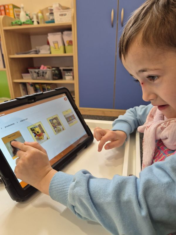 Dziecko w niebieskiej bluzie używa tabletu z aplikacją edukacyjną w kolorowym pokoju.
