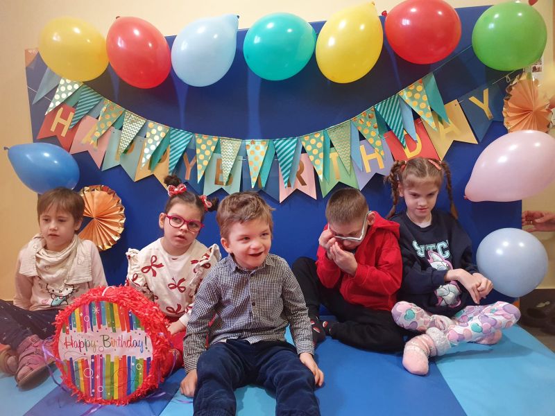 Dzieci siedzą na tle urodzinowych dekoracji z balonami i napisem Happy Birthday.
