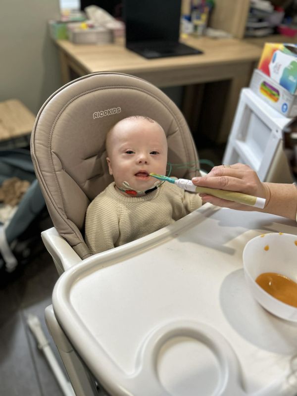 Dziecko w krzesełku do karmienia, karmione łyżeczką przez dorosłego. Obok miska z puree.