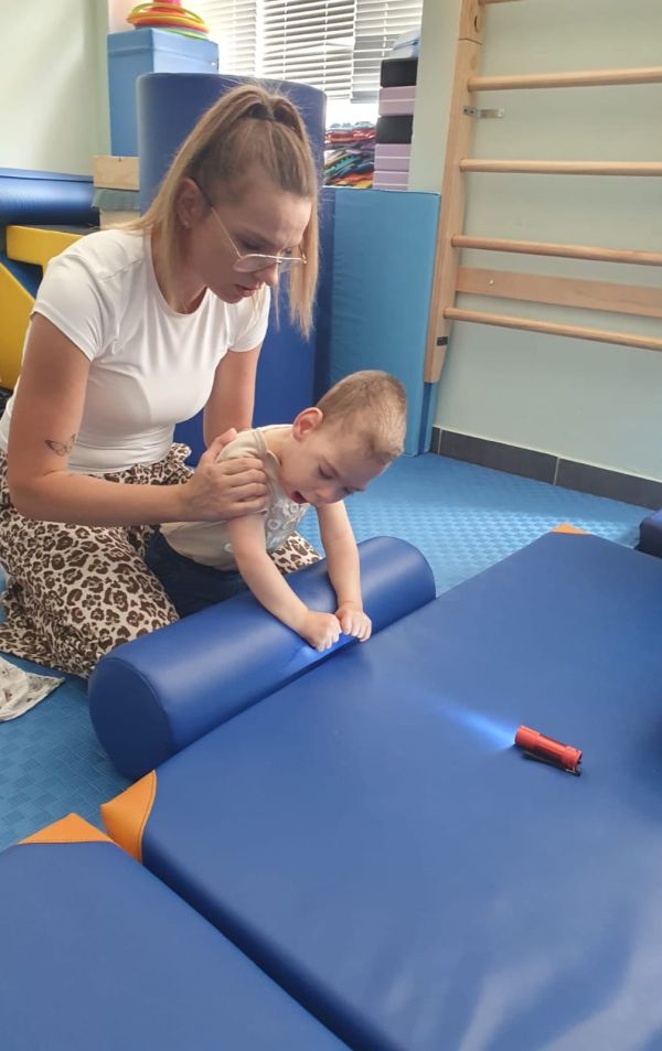 Osoba pomaga dziecku przytulonemu do niebieskiej maty, w sali do ćwiczeń z drabinkami na ścianie.